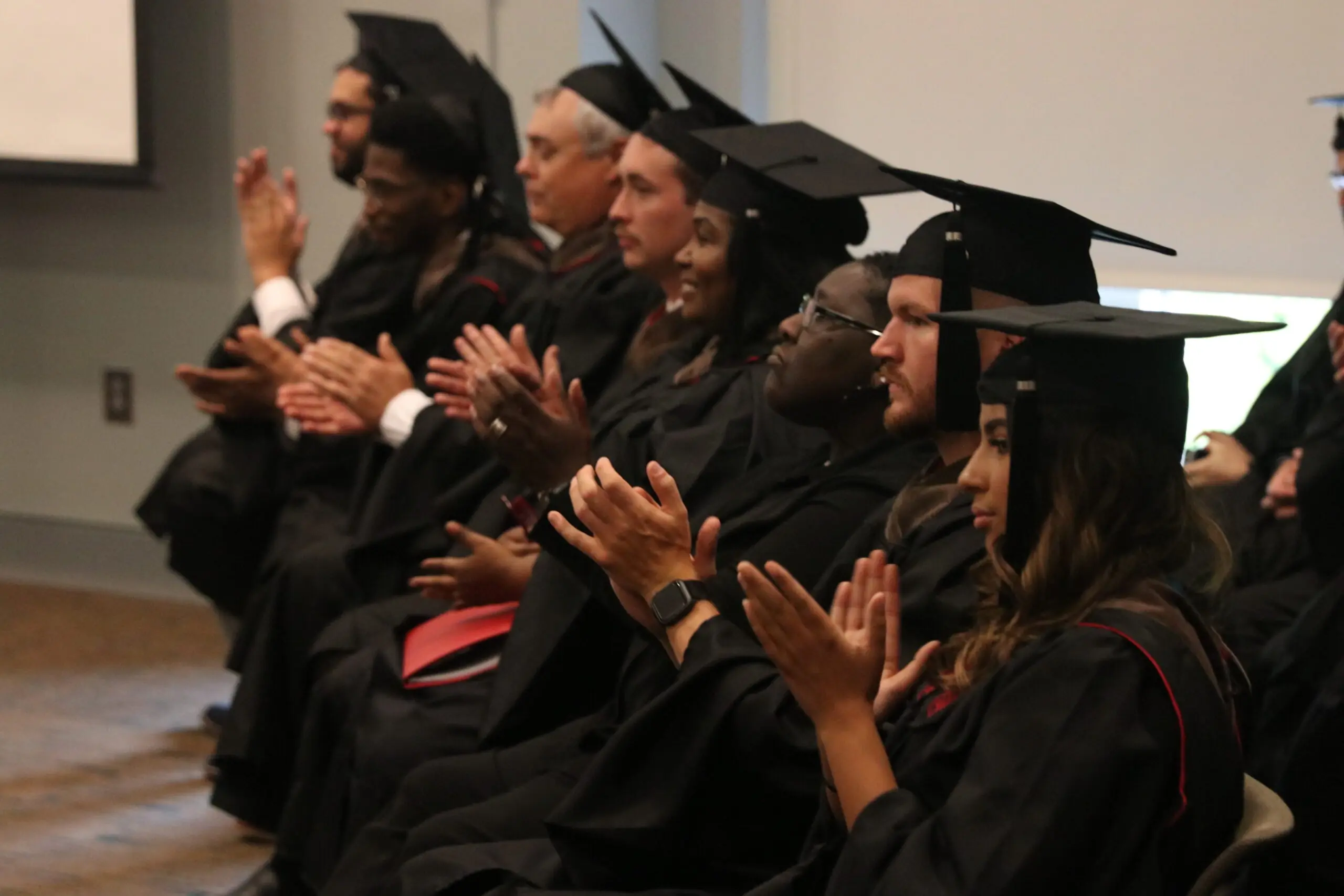A group of people in graduation regalia sit in a row of chairs applauding.