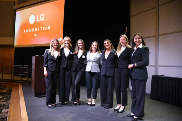 Group of 7 students pose for a picture on a stage with a screen in the background that says LG.