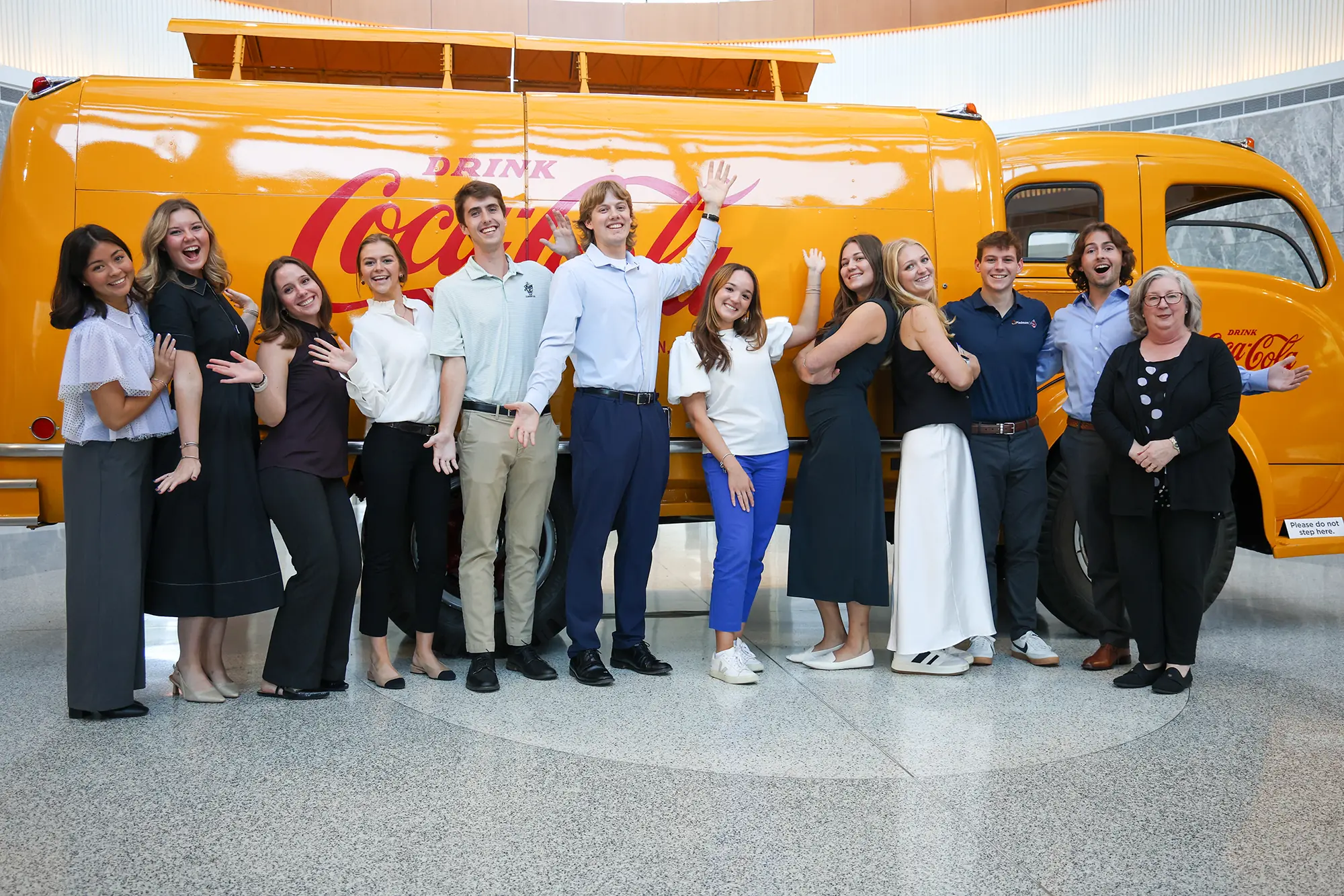 A group of Talking Dog students raise their arms, smile and pose for a picture inside the Coke museum. They stand in front of a old-fashioned yellow truck with a red Coca Cola emblem on the side.