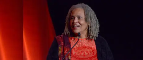 A picture of Charlayne Hunter Gault speaking at a formal even with red and black piping in the background.