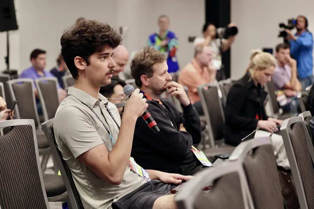 Close up profile of a student holding a microphone asking a question in a group of people sitting down. Cameras are out of focus in the background.