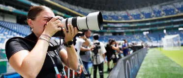 A female photographer has a long-lens camera and takes a pictures with a large stadium and field in the background.