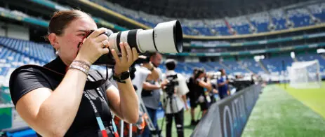 A female photographer has a long-lens camera and takes a pictures with a large stadium and field in the background.