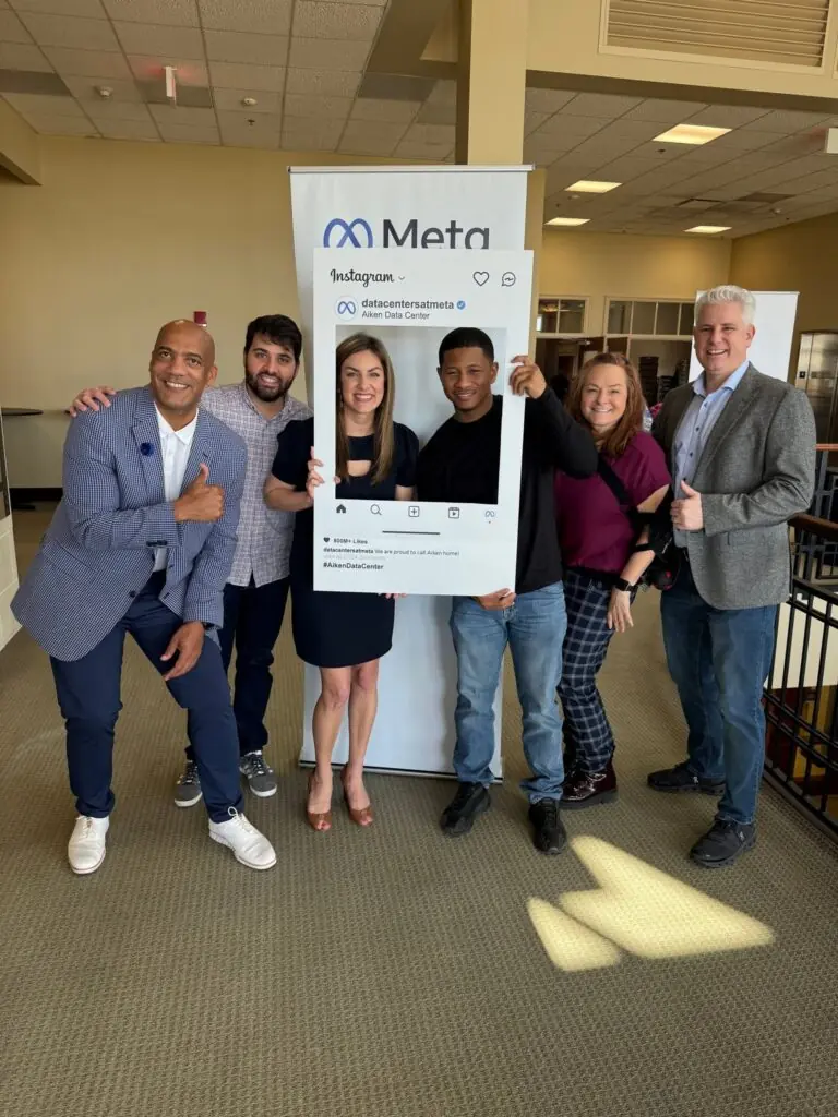 A group of people, four men and two women, pose for a picture in professional clothing, with one woman and one man holding a sign that looks like an Instagram post. 