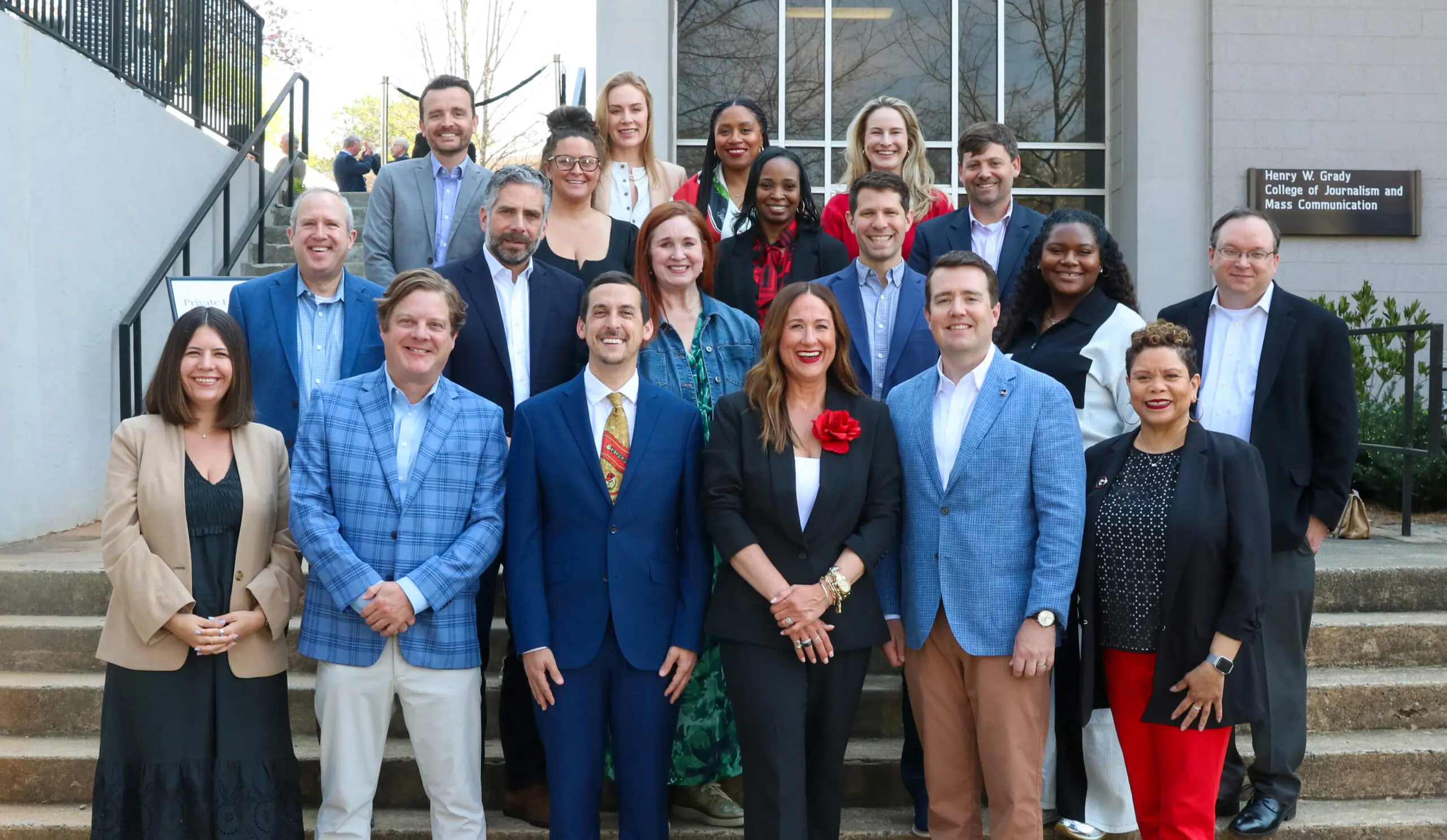 A group of about 25 people pose for a picture on a set of stairs. In the background is a sign that reads Henry W. Grady College of Journalism and Mass Communication. 