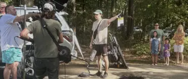 A man holds a film slate as a camera crew gets ready to film a family, seen in the background.