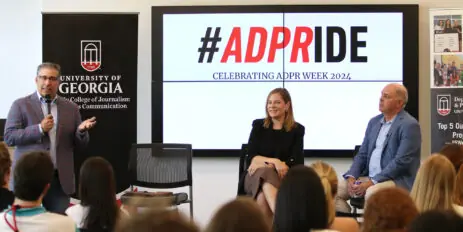 A man holding a microphone and a man and woman sitting talk with a large group of students. A digital projection in the background says #ADPRIDE.
