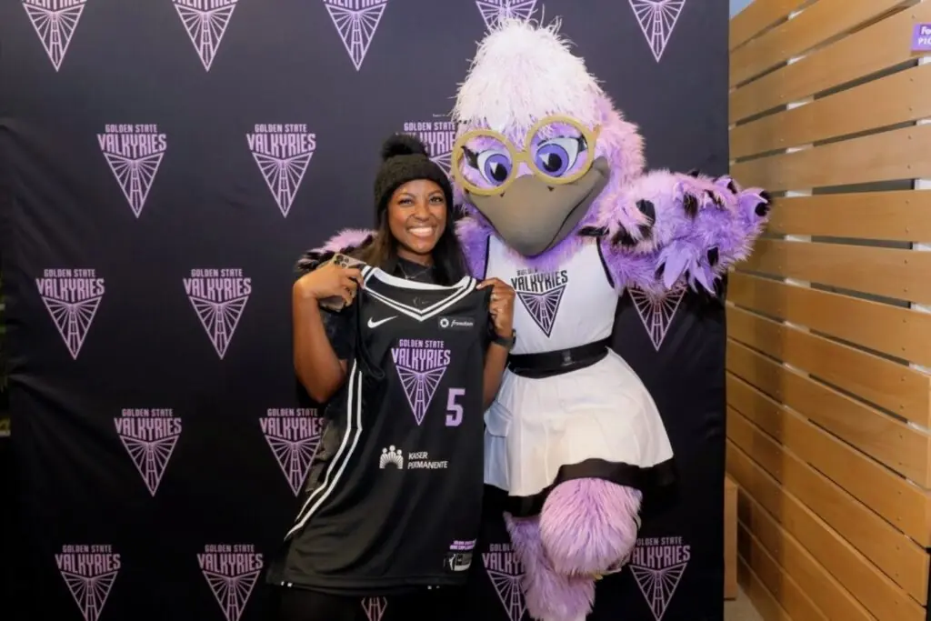 Renee Fuller stands in front of a Golden State Valkyries backdrop next to the Golden State Valkyries mascot, a large purple bird.