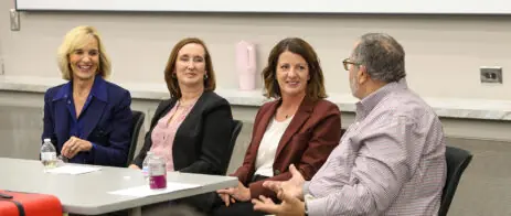 Three women dressed in business attire sitting on one side of a table look toward Dean Davis who has his back toward the camera.