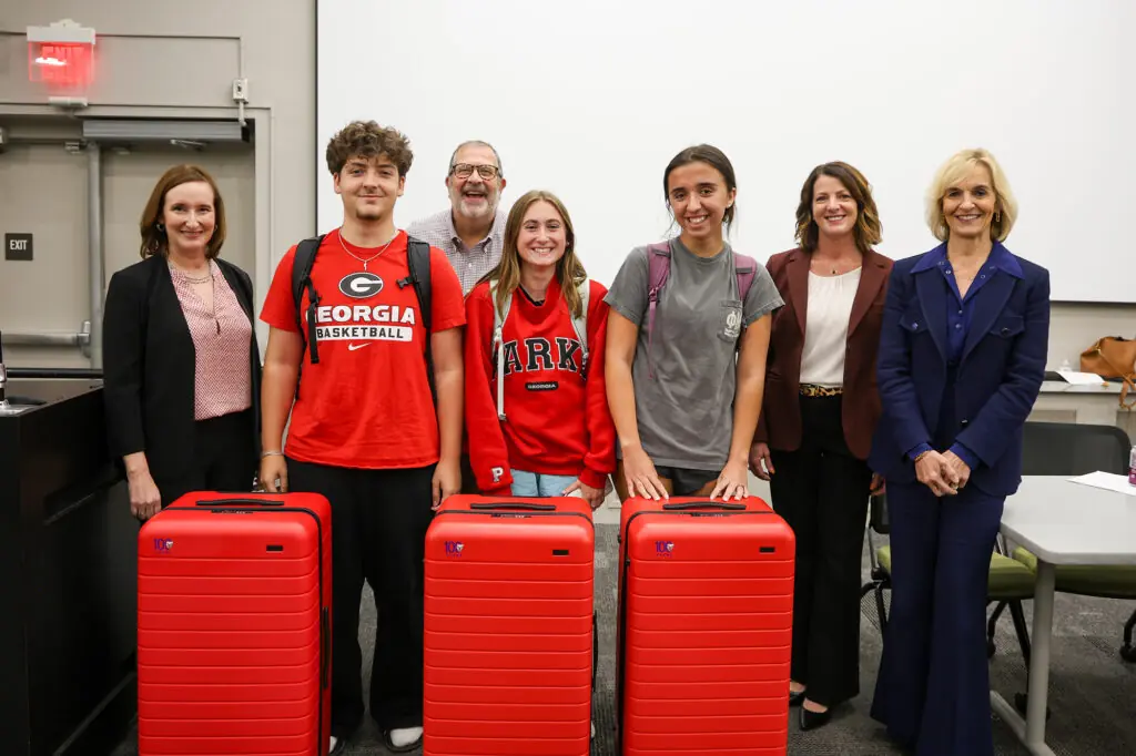 Three students with three large, red suitcases in front of them pose with four adults dressed in business casual attire.