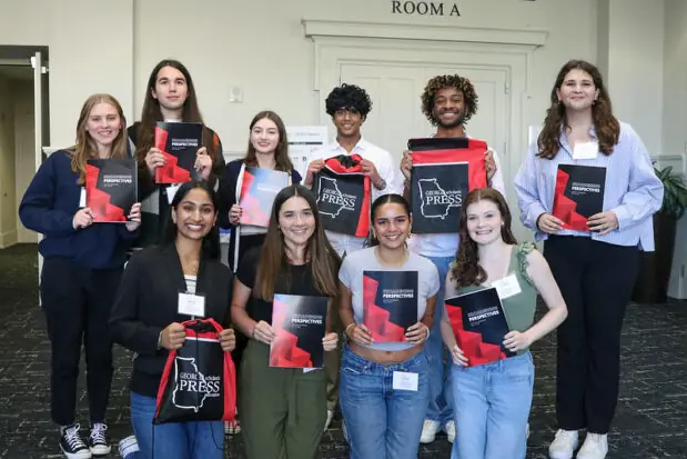 A group of eight smiling young adults stand indoors, holding up black and red drawstring bags labeled Georgia Scholastic Press. Two people wear blue shirts that read, We have issues.