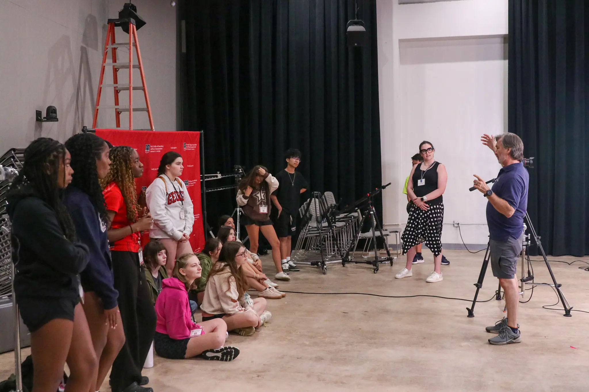 A group of students listens to an instructor gesturing while standing in a studio with black curtains, a camera on a tripod, and a red ladder in the background. Some students are seated on the floor, others stand.