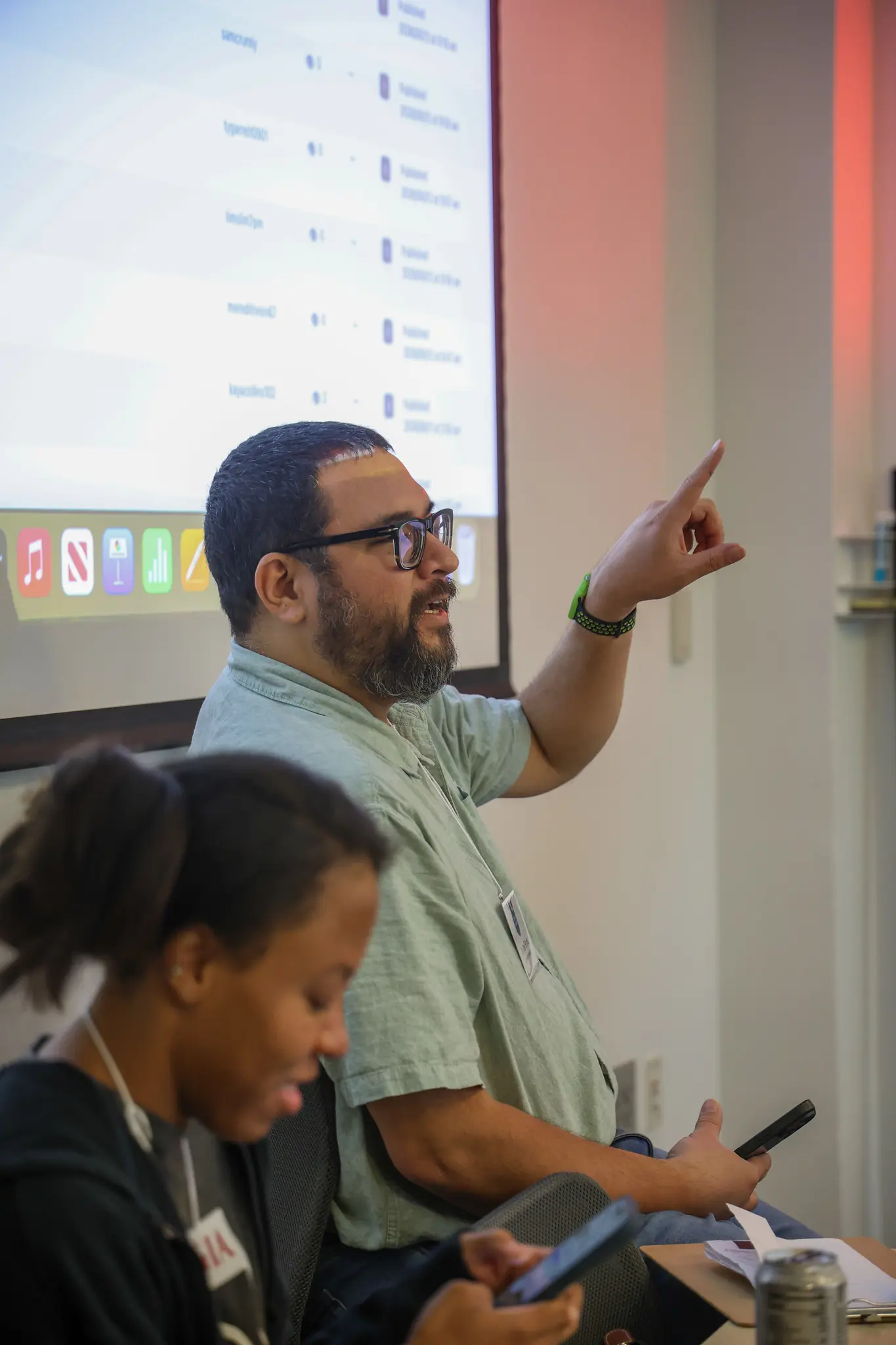 A man with glasses raises his hand while speaking in a meeting room; a woman in the foreground looks down, and a projection screen with app icons is visible behind them.