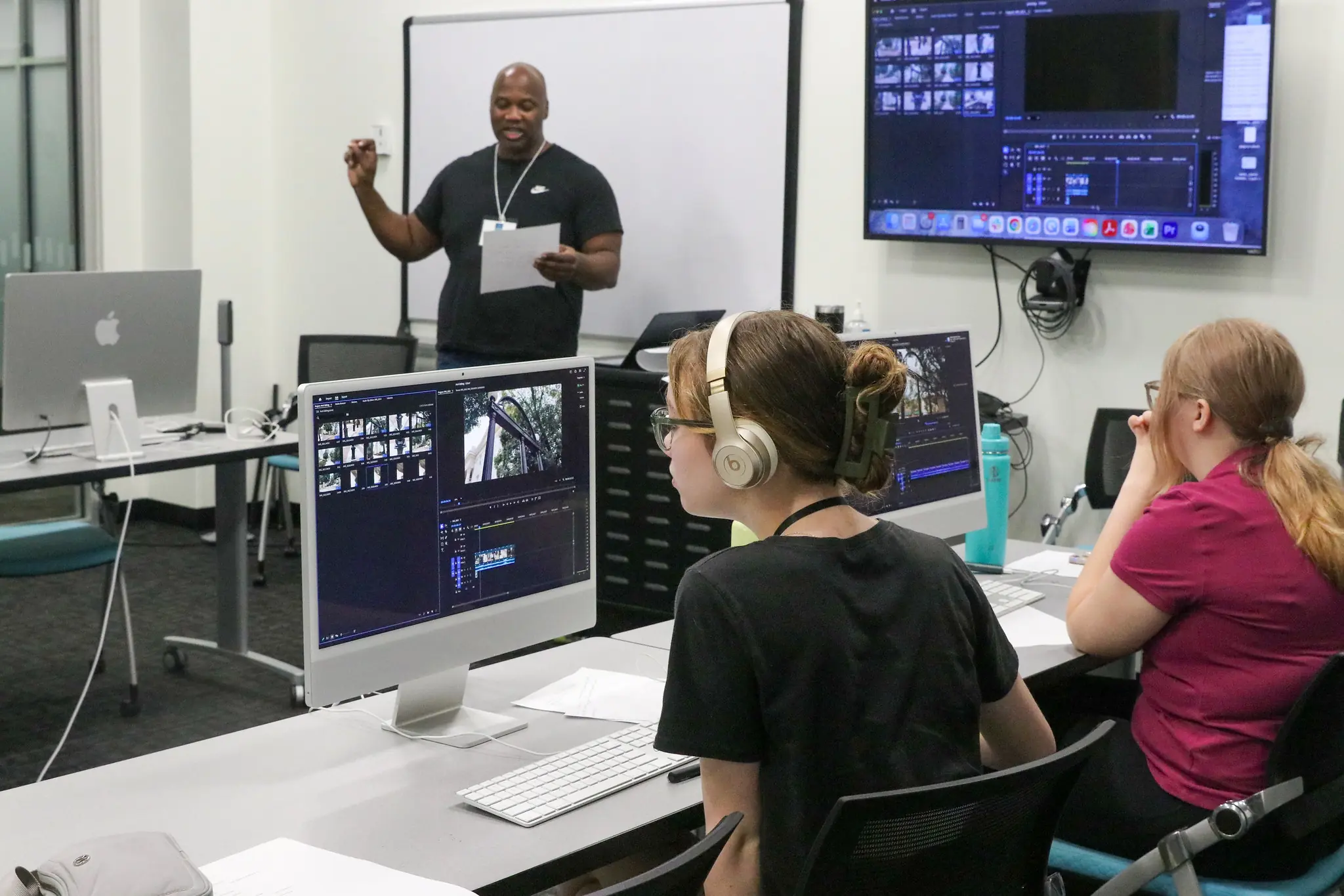 Three people sit editing videos on computers, wearing headphones, while an instructor stands at the front holding paper, speaking. Large screens display video editing software. Classroom setting.