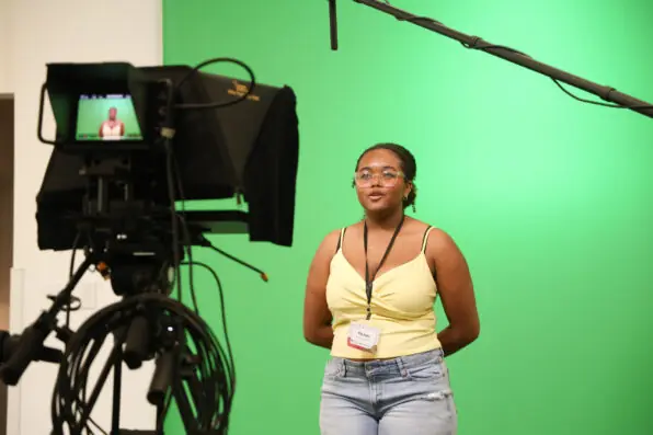 A teenager stands in front of a green screen, speaking into a camera with a microphone overhead. They wear glasses, a yellow top, jeans, and a name badge. A monitor shows their image as they record.