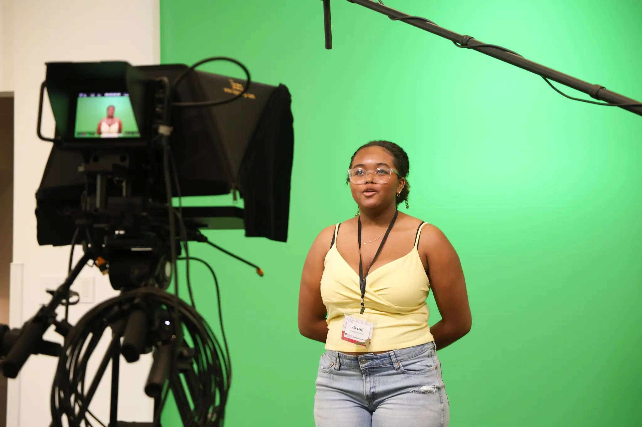 A teenager stands in front of a green screen, speaking into a camera with a microphone overhead. They wear glasses, a yellow top, jeans, and a name badge. A monitor shows their image as they record.